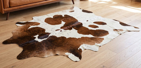 A natural cowhide area rug with a brown and white mottled pattern laid out on a light wood floor in a modern living room setting with a brown leather sofa.