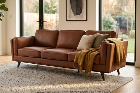 A mid-century modern brown leather sofa in a bright, modern living room with large windows, a potted fiddle-leaf fig, and a textured area rug.