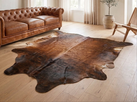 Brown cowhide rug with dark markings on a wooden floor beside a cognac leather Chesterfield sofa.