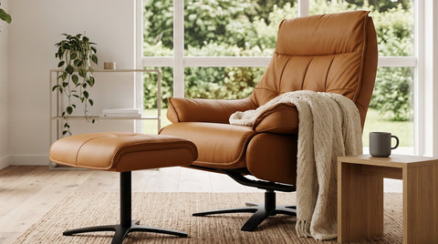 A tan leather recliner chair with an adjustable ottoman and a draped cream throw blanket, styled in a bright, modern living room with large windows, a potted fiddle-leaf fig, and a wooden side table.