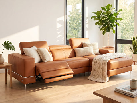 A modern sectional sofa in warm tan leather, styled with cream throw pillows and a cream knit blanket in a bright, sunlit living room with large windows and potted plants.