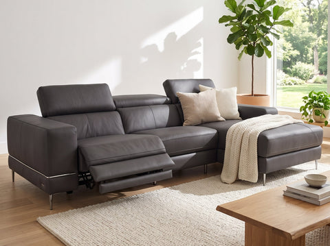 A dark gray leather sectional sofa with a chaise lounge and extended footrest, styled in a bright, modern living room with a wooden coffee table, textured rug, and potted plants.