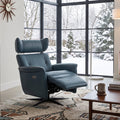 A mid-century modern living room featuring a blue leather recliner chair positioned in front of large black-framed windows looking out onto a snowy winter landscape. The room is styled with a mid-century wooden bookcase, starburst wall clocks, a patterned rug, and a coffee table.