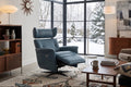 A mid-century modern living room featuring a blue leather recliner chair positioned in front of large black-framed windows looking out onto a snowy winter landscape. The room is styled with a mid-century wooden bookcase, starburst wall clocks, a patterned rug, and a coffee table.