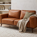 A warm, modern living room featuring a brown leather sofa, a woven wood and cane armchair, a large white textured area rug, and a tall floor lamp, set against cream plaster walls and a skylight.